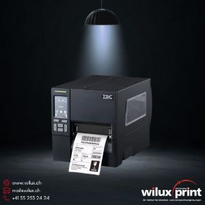 Schwarzer industrieller Etikettendrucker der Marke TSC, beleuchtet von einem Spotlicht, der ein gedrucktes Barcode-Etikett ausgibt – WILUX PRINT Blog