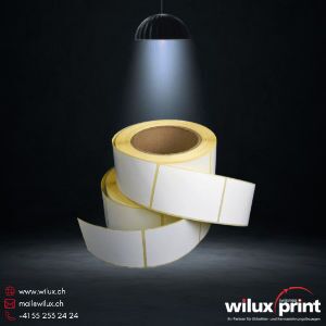 Zwei aufgerollte weisse Papieretikettenrollen auf dunklem Hintergrund, beleuchtet von einem einzelnen Spotlicht, für industrielle und logistische Anwendungen – WILUX PRINT Blog.